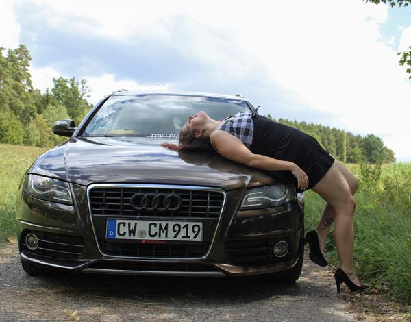 Frau posiert auf der Motorhaube eines Autos vor grünem Feld